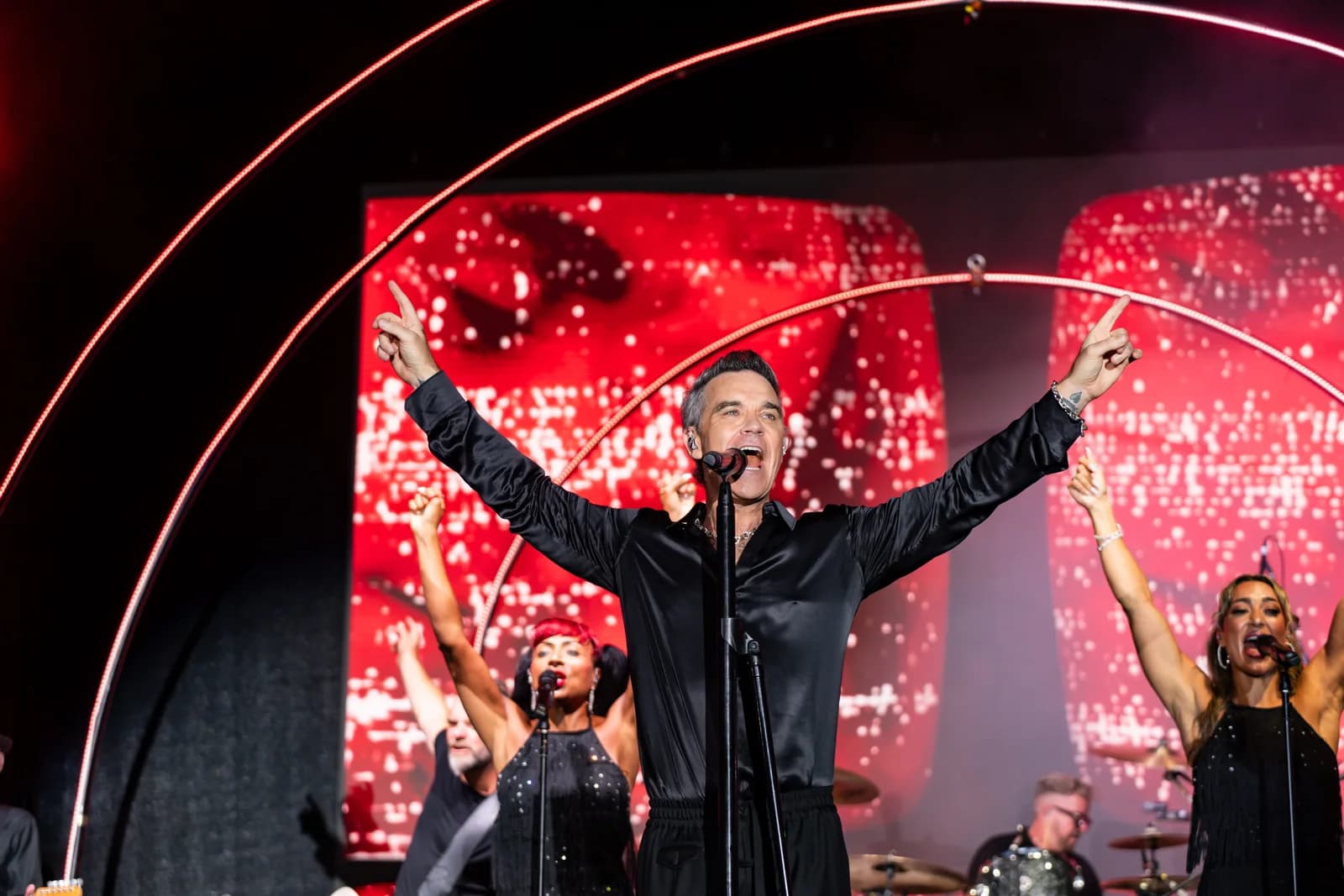 Robbie Williams performs with arms raised on the BIG ART Festival stage in Porto Cervo, backed by red lighting