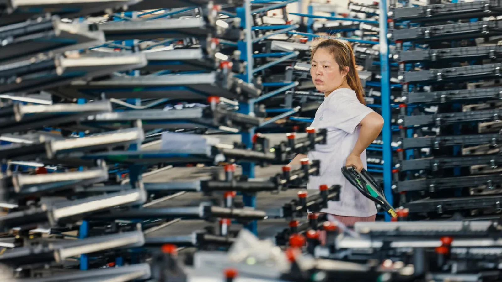 A worker stands among towering racks of stacked electronic components on a busy factory production line